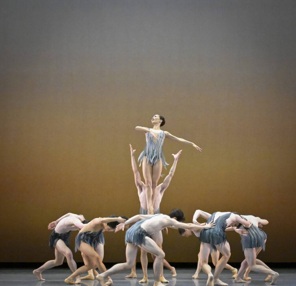 A dancer hovers in the air in sous-sus looking up towards the left diagonal with her mouth open. her left arm bends at the elbow with her hand tilted down towards her chest with her right arm extended to the right. She wears a grey tank leotard embellished with featherlike strips of grey fabric. circling beneath her are a circle of dancers in similar costumes hunched over in counterclockwise-facing plie fourth position connected together with their curved arms.