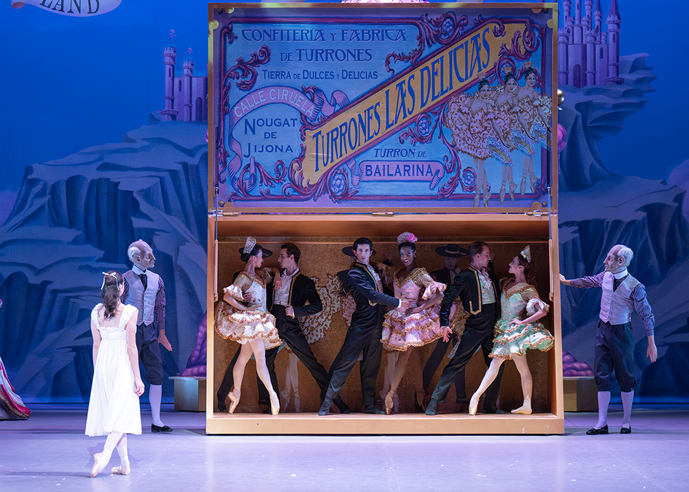 Three couples pose inside of an open horizontal wooden box. The male dancers wear black suits with gold trimming and the females wear colorful layered pancake tutus.