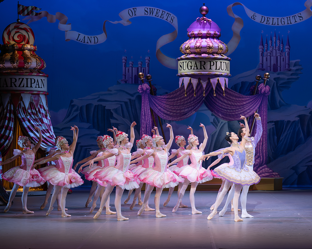 The Sugar Plum Fairy and Sugar Plum cavaliar stand facing the right leading a group of dancers in pink behind them. They all stand with their left leg and left arm extended to the back with their right arm arched above their head.