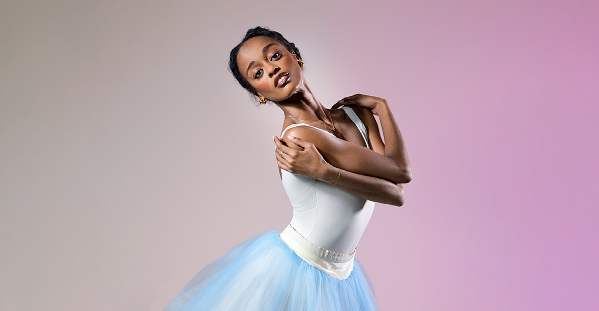India Bradley, wearing a white leotard and blue tutu, is shown from the thighs up. She crosses her arms over her chest, holding her shoulders and looks toward the camera with a small smile, arching her upper body back slightly.