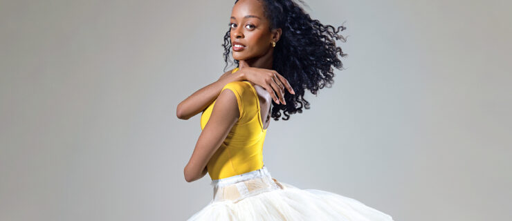 India Bradley, wearing a bright yellow leotard and white practice tutu, is shown from the thighs up in profile. She crosses her arms, placing her right hand over her left shoulder, and looks out towards th4 camera with a small smile.