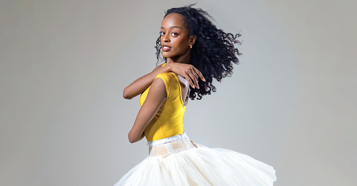 India Bradley, wearing a bright yellow leotard and white practice tutu, is shown from the thighs up in profile. She crosses her arms, placing her right hand over her left shoulder, and looks out towards th4 camera with a small smile.