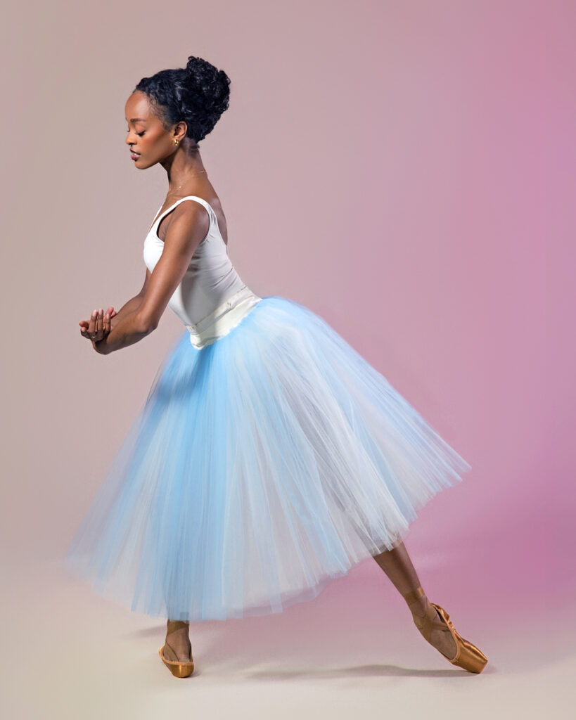 India Bradley wears a white leotard and light blue romantic tutu. She poses in profile, facing stage right, in tendu derriere with her left leg back. She crosses her wrists in front of her and looks down at her hands.