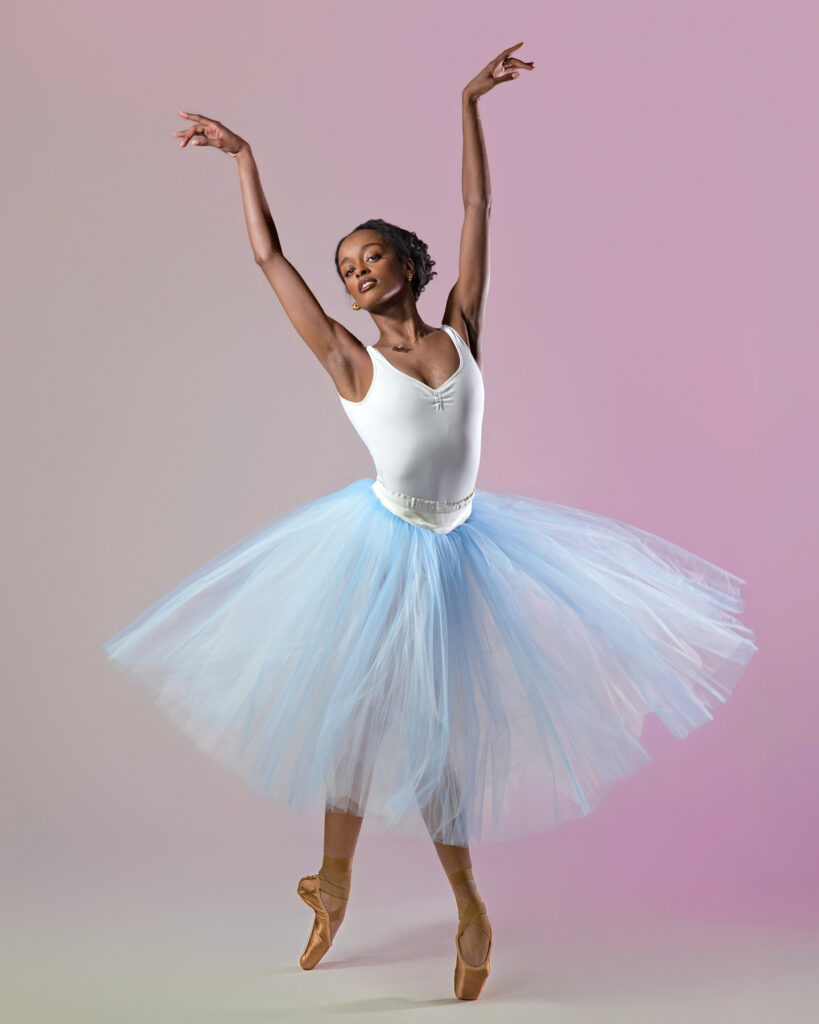 India Bradley, wearing a white tank leotard, light blue romantic tutu, and brown pointe shoes, poses in fourth position croisé on pointe. She lifts both arms up above her head and tilts her head slightly to the right. She poses in front of a gray backdrop with a pink ombre effect.