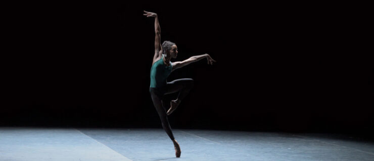 Onstage in stark lighting, Precious Adams does a leaning retiré en pointe, arms extended on the diagonals in a wide tilted "V." She wears a teal leotard and black tights.