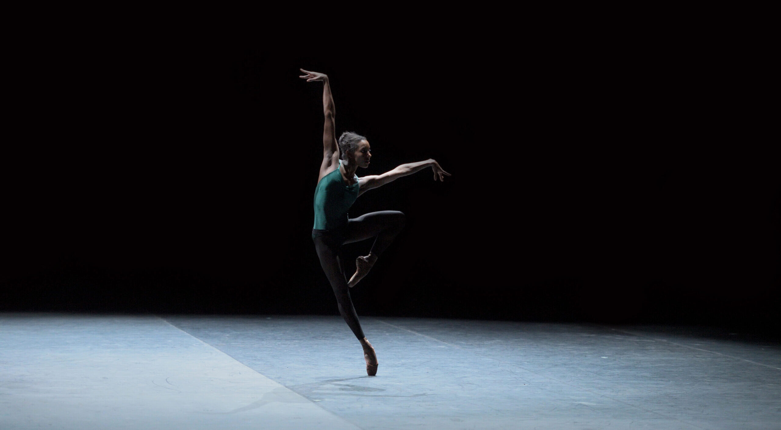 Onstage in stark lighting, Precious Adams does a leaning retiré en pointe, arms extended on the diagonals in a wide tilted "V." She wears a teal leotard and black tights.