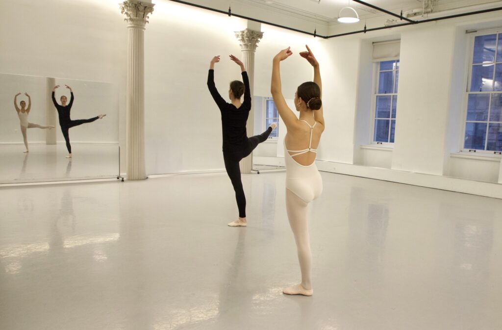 In a light-colored ballet studio, Natalia Boesch demonstrates a developpé a la seconde as a student in a white leotard and tights follows her movement. Their reflections are visible in a large mirror at the front of the room.