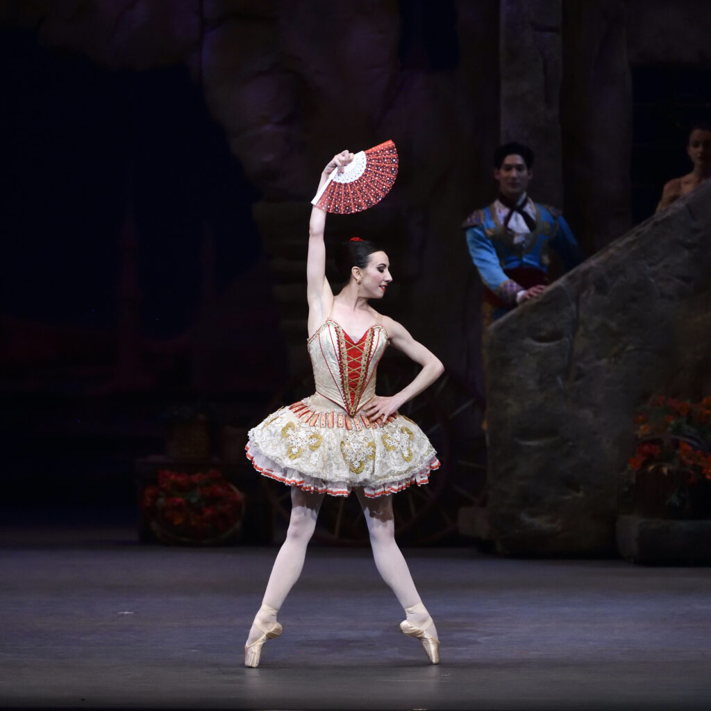 Boylston poses en pointe in échappé second while looking downward to the right, placing her right hand on her hip with her left arm holding an open downward-facing red and gold fan above her head. She wears a matching gold tutu with red and white detailing and a rose hairpiece tucked into her french twist.