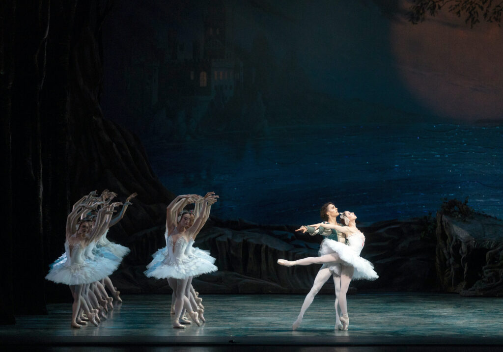 Boylston performs the role of Odette partnering with Camargo as Prince Siegfried. She wears a white pancake tutu and feathered headpiece while balancing on her right leg in right-facing arabesque, arching backwards with her arms extended behind her. Camargo supports her left arm with his left arm extended to the side underneath hers and his chest pressed against her back while mirroring her pose with his right leg extended in a back tendu. He wears white tights and a long-sleeve green velvet top featuring cream and gold accents. Behind them to the far left, fourteen swan dancers wearing white feathered pancake tutus and matching feathered headpieces stand in two front-facing lines of seven. They pose with their left legs behind them in B-plus and their arms curved to the right above their head while looking towards a downward right diagonal.