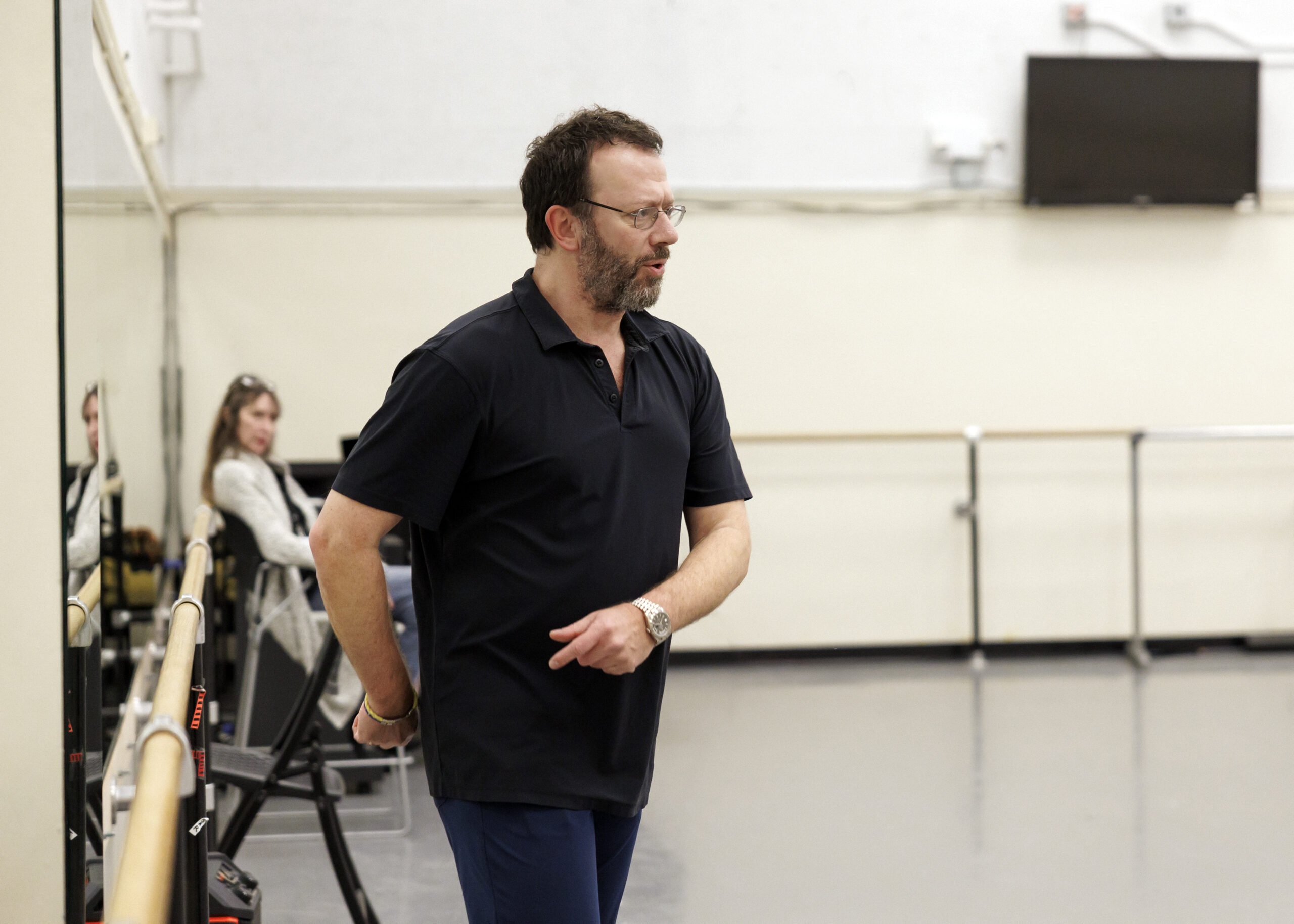 In a bright dance studio, Alexei Ratmansky addresses a room of dancers (not shown) and gently wraps his arms around his right hip.