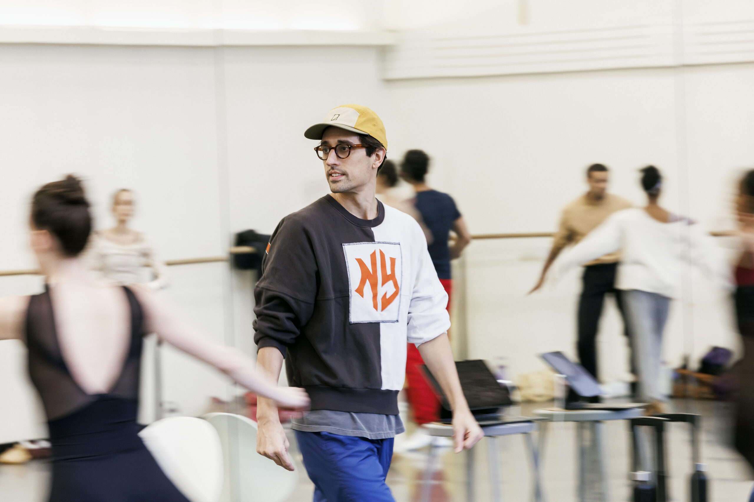 In a large ballet studio, Justin Peck watches as dancers run around him with their arms extended gently to either side. 