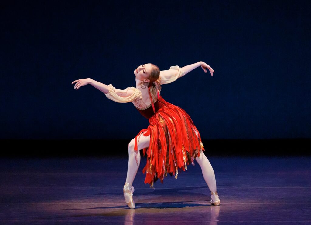 Dominika Afanasenkov is spotlit, front leg in forced arch on pointe. Her torso is twisted to face upstage as she reaches forward, arms a sinuous curve that matches her legs. She wears a dress with voluminous white sleeves, a dark bodice, and a layered red skirt over pink tights and pointe shoes.
