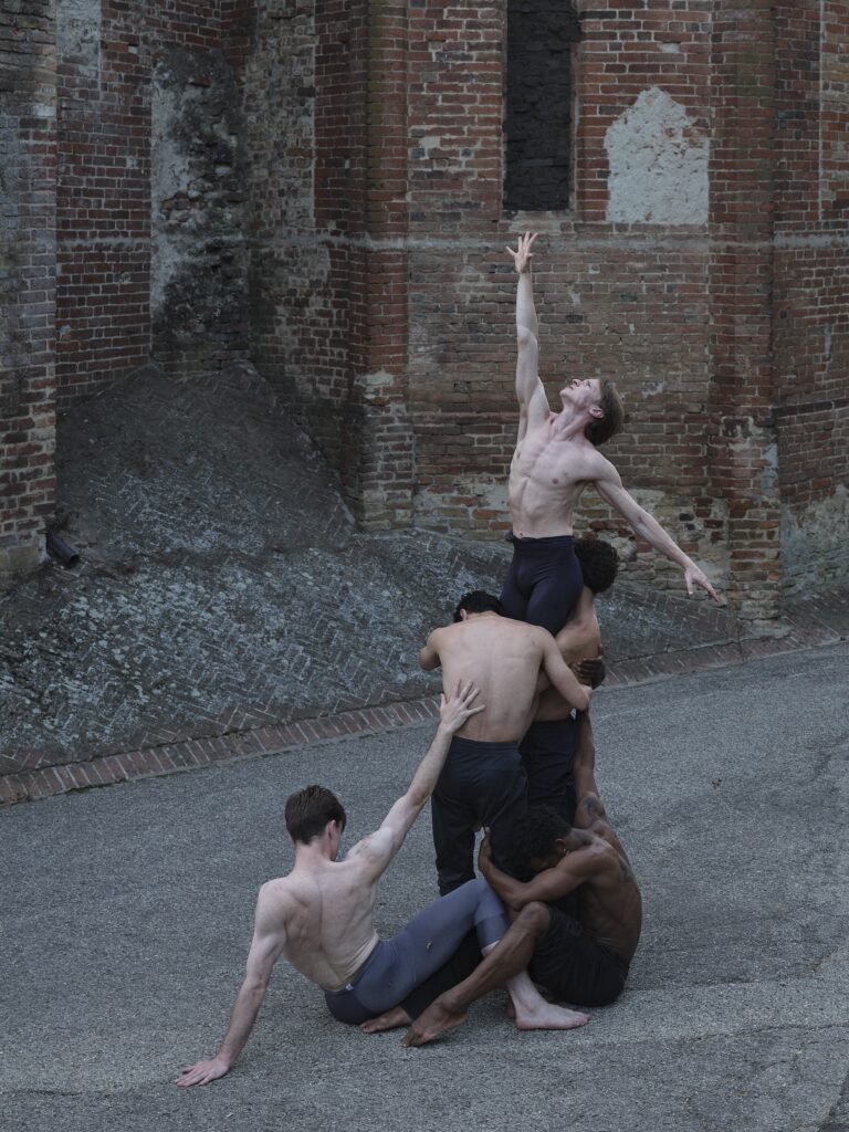 A group of male dancers in dark tights are arranged in a tight, huddled formation, with one dancer being lifted above the rest, reaching his arm high, and two others sittiing on the ground. They pose outdoors on the grass near a brick building at dusk.