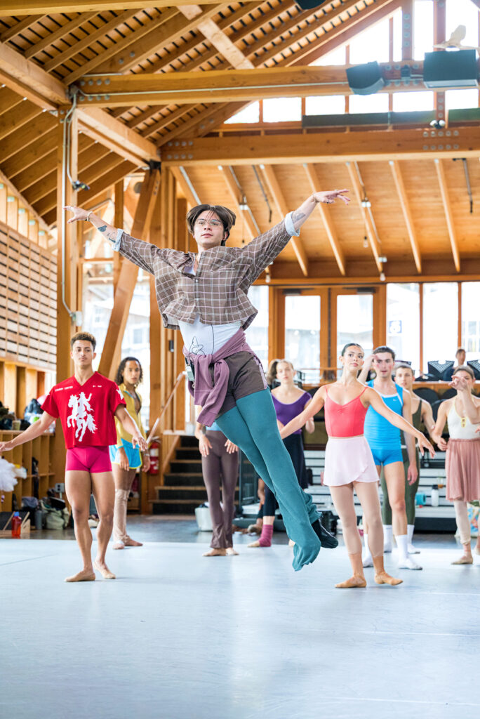 Durante Verzola, wearing colorful rehearsal gear and glasses, demonstrates an assemblé for a studio full of ballet students.
