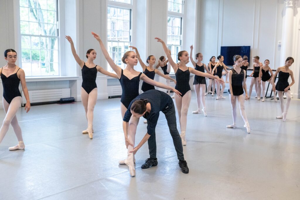 In a large, well-lit dance studio, a ballet teacher in practice clothes reaches down to adjust the lower leg of a young dancer in tendue devant croisé during a class. All of the girls wear black leotards, pink tights, and pink pointe shoes, and a few wear short black ballet skirts.