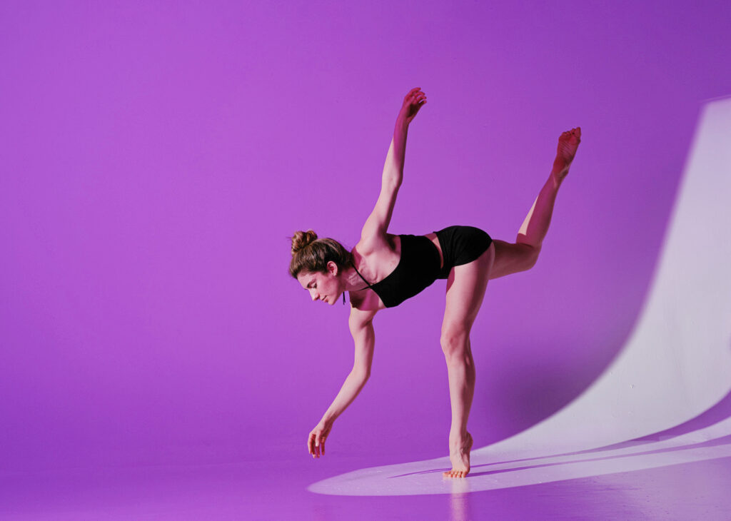 Juliette Ochoa balances on forced arch, torso parallel to the floor, back leg raised in a parallel attitude. One hand bends gracefully toward the floor, while the other rises behind her. She wears a fitted black tank top and black shorts; the background is a bright purple.