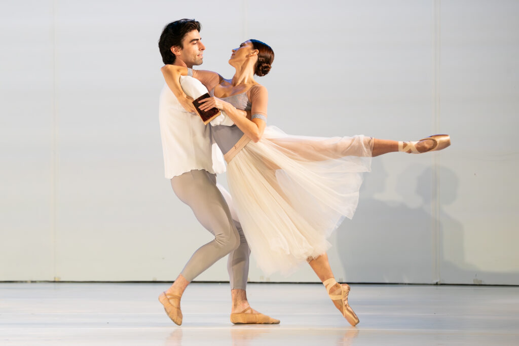 In a bright, light-colored space, two dancers perform a pas de deux in neutral-colored costumed. A ballerina leans into her partner in an arabesque position, holding a book and hooking one arm around her partner's neck. He walks backward, dragging her with him as they look into each other's eyes.