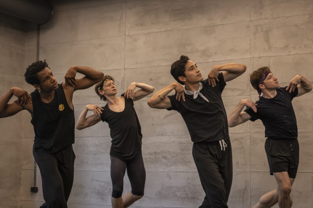 Osiel Guneo, Daniil Simkin, Jeffrey Cirio, and Alban Lendorf rehearse in a dance studio with cement walls. They stand in a staggered line and place their hands on their shoulders, their left elbow slightly up, as they walk towards their left. They wear practice clothes in dark colors.