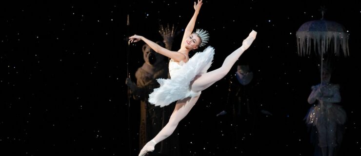 Tyler Donatelli, wearing a white tutu, pink tights, pointe shoes, and a spiky silver crown, jumps up with her arms in third arabesque and her back, left leg high in attitude behind her. She looks out towards the audience with a joyful smile, and snow falls around her and onto the stage.