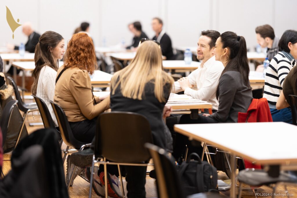 A teenage girl and two adults sit at a table across from a man and woman and talk in a large meeting room. Other tables with people sitting at them surround them.