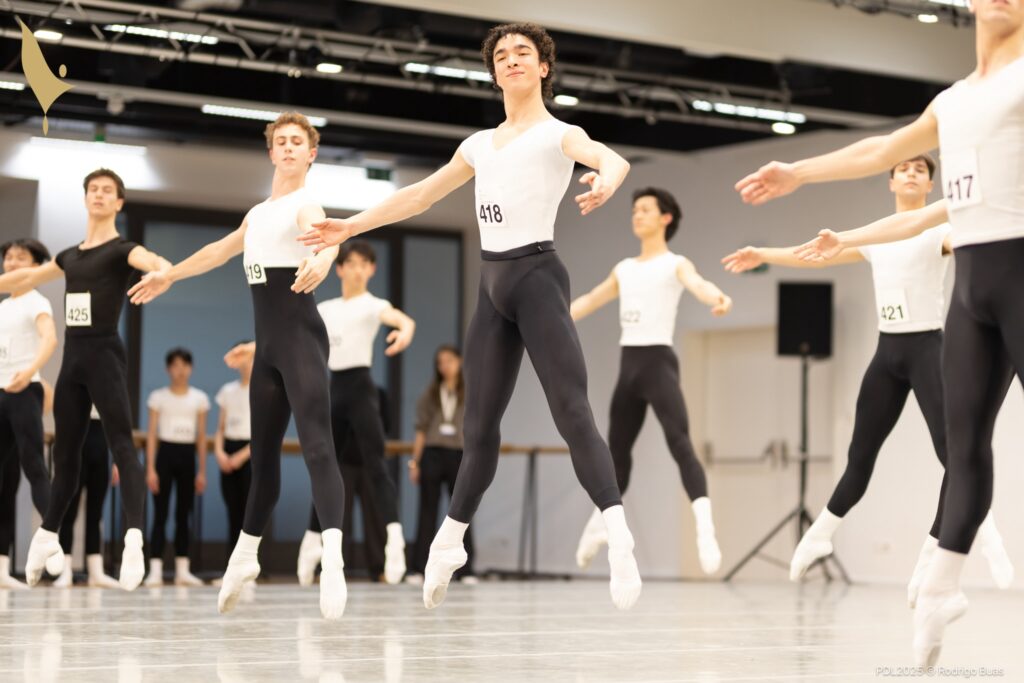 Two lines of teenage ballet students do échappé sauté during a ballet class in a dance studio. They all wear black tights, white socks, and white ballet slippers. Most also wear white t-shirts, save for one dancer in a black t-shirt.
