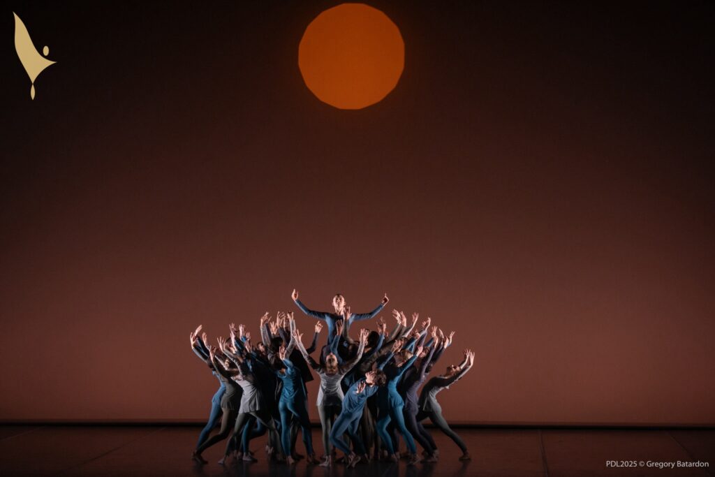A group of young dancers in blue and gray costumes create a circular formation, lunging towards the center with their arms raised and backs slightly arched. A dancer in the middle faces the audience and is lifted above the others. The backdrop is a dark brown, with an orange sun in the center, towards the top.