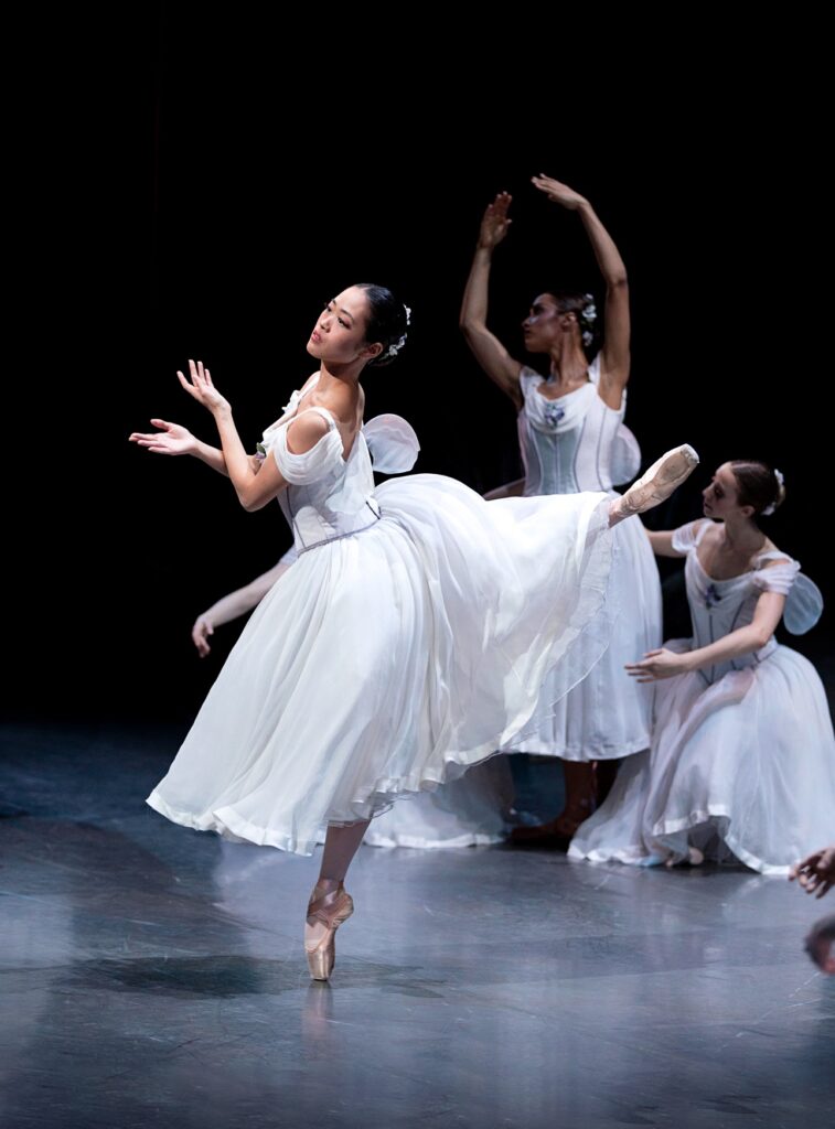 Yuki Takahashi is photographed from the wings as she balances in attitude back, forearms crossed just in front of her torso. She is costumed in a white romantic tutu with small wings, pink tights, and pointe shoes.