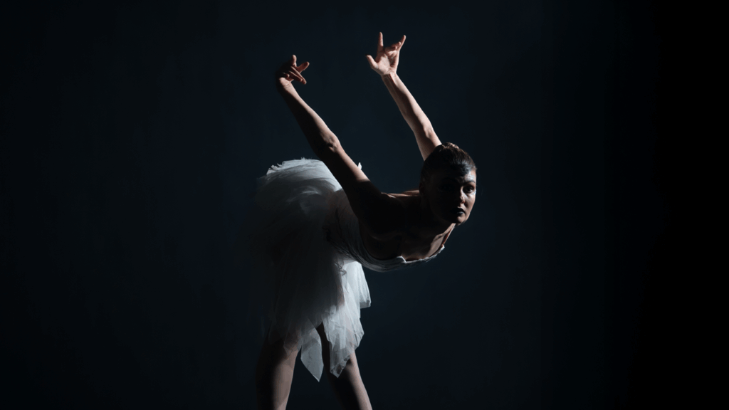 In front of a black background, a ballet dancer in stark lighting poses on pointe with her arms reaching up and back, like wings, as she leans forward. She wears a white dress with a distressed bottom that hits just around her knees.