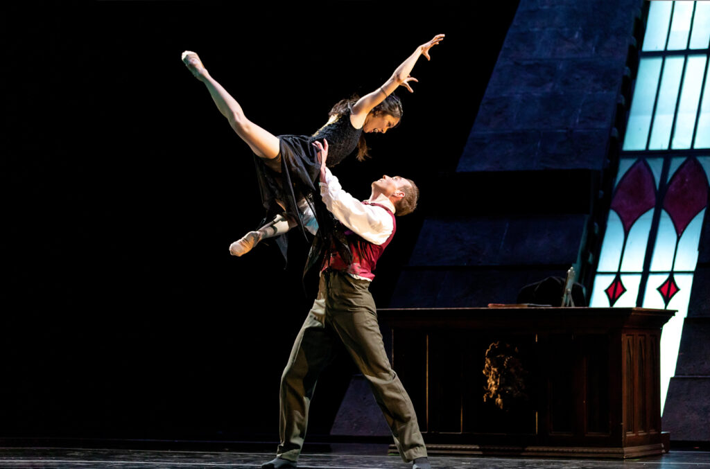 Sumi Ichikawa hisses dramatically as she is held overhead in a lift. Her legs are bent in double attitude, and her arms are raised overhead, fingers flexing into claws. Her hair is loose to her shoulders, and she is costumed in black. The set features a stained glass window and a wooden desk.