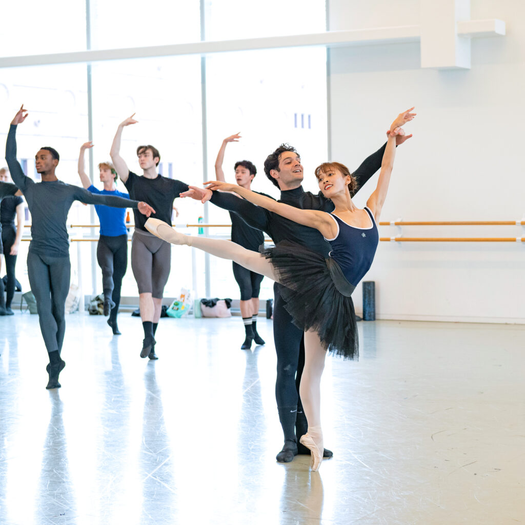 In a brightly lit studio, Sayako Toku balances in a high first arabesque, glancing back over her shoulder toward her partner, who lightly holds the wrist of her front arm.