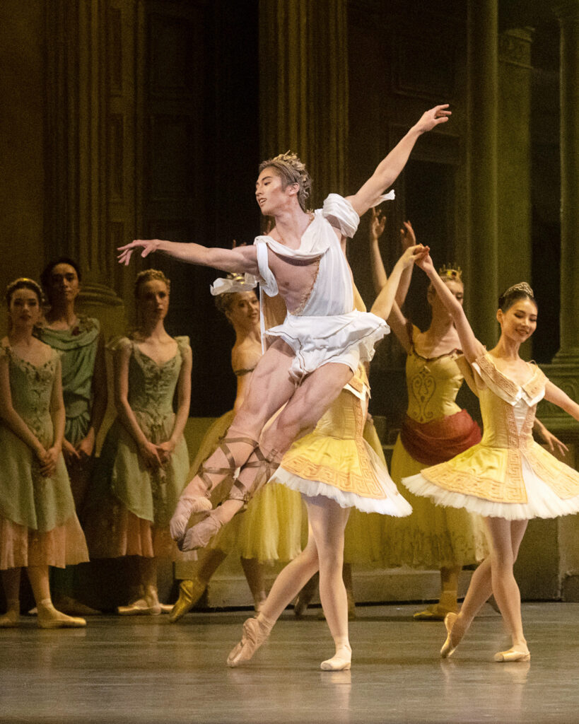 Takumi Miyake is captured midair in brissé front. His costume evokes a Grecian toga and sandals.