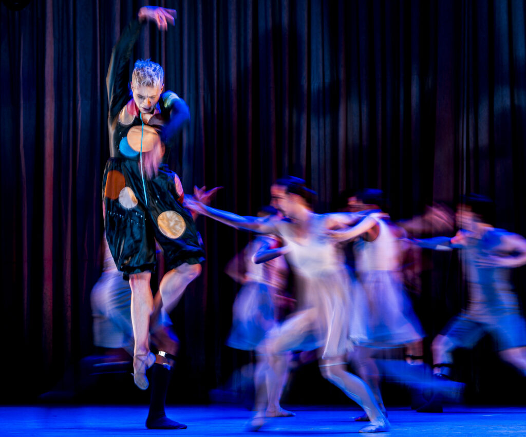 On a dark stage, a group of dancers move behind a solo dancer in front, who jumps with one leg in retiré derriere and arms in high fifth. They wear circus-style costumes, and the image is blurred with movement.