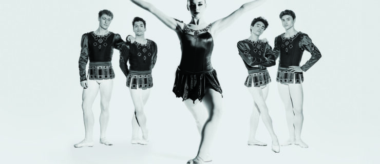 A silver-tone image of five dancers in "Rubies," with four men standing casually in the back as a woman, in center, leans forward with one foot propped on pointe. She flexed her hands and reaches her arms up on a "V" shape as she looks into the camera.