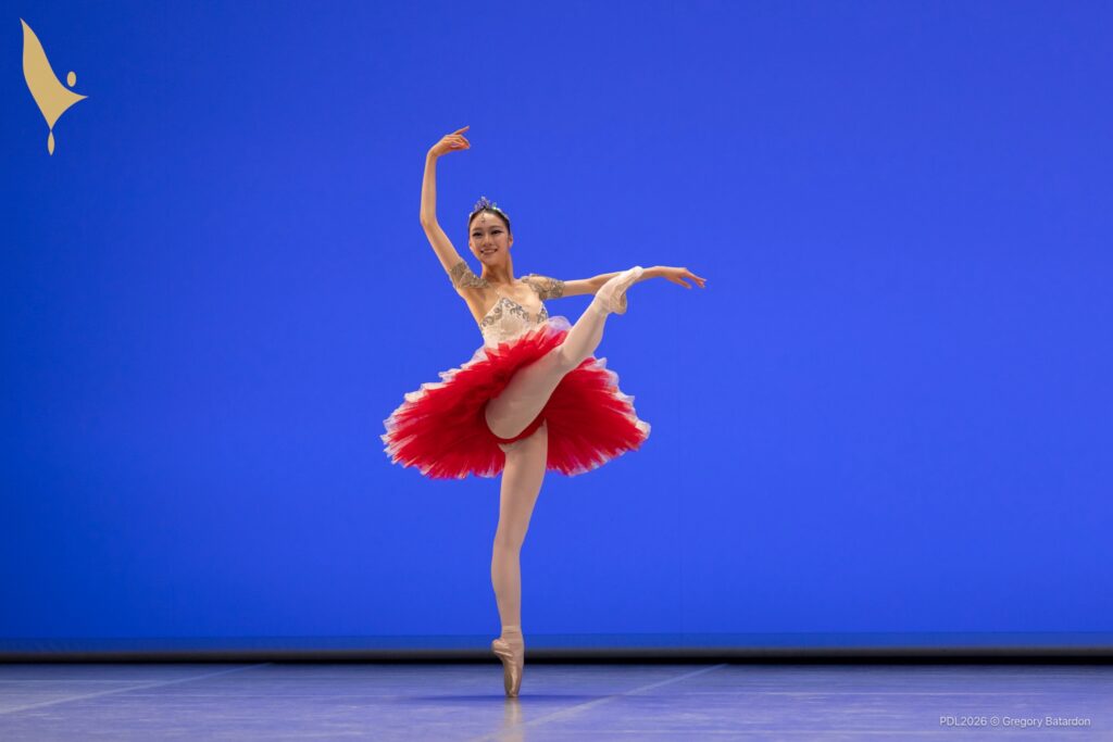 A teenage ballerina does a développé croise devant with her right leg on pointe during a performance. She wears a white tutu with layers of red underneath and a tiara. She looks out to the audience and smiles.