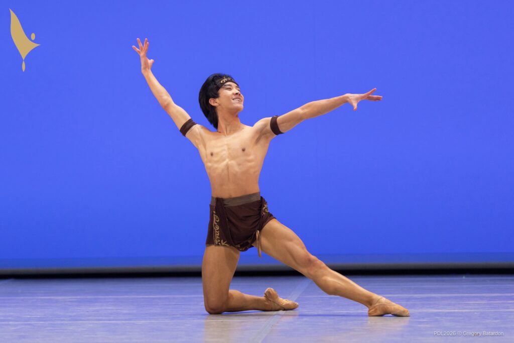 A teenage ballet dancer kneels on his right knee in an open position, lifting his right arm and looking out triumphantly over his extended left hand. He wears brown shorts, brown arm bands, and a brown bandana.
