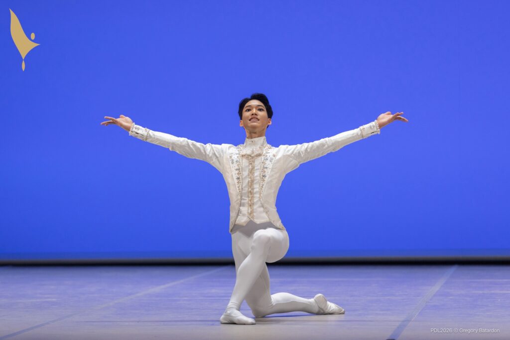 A teenage ballet dancer in a white tunic and tights kneels onto his right knee, facing directly front. He opens his arms wide and smiles out to the audience.