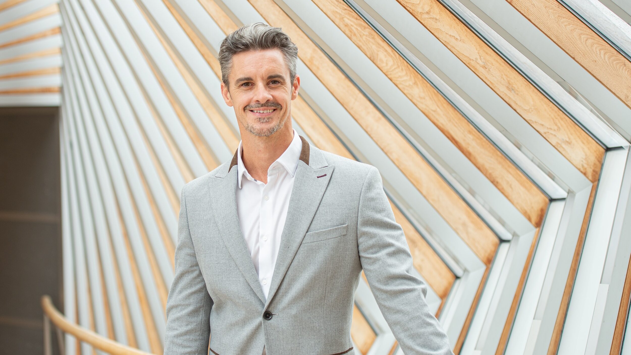 Iain Mackay is shown from the waist up. He wears a light gray sport coat over a white button down shirt and smiles towards the camera. He stands in a hallway with curved walls featuring panels with a modern design.