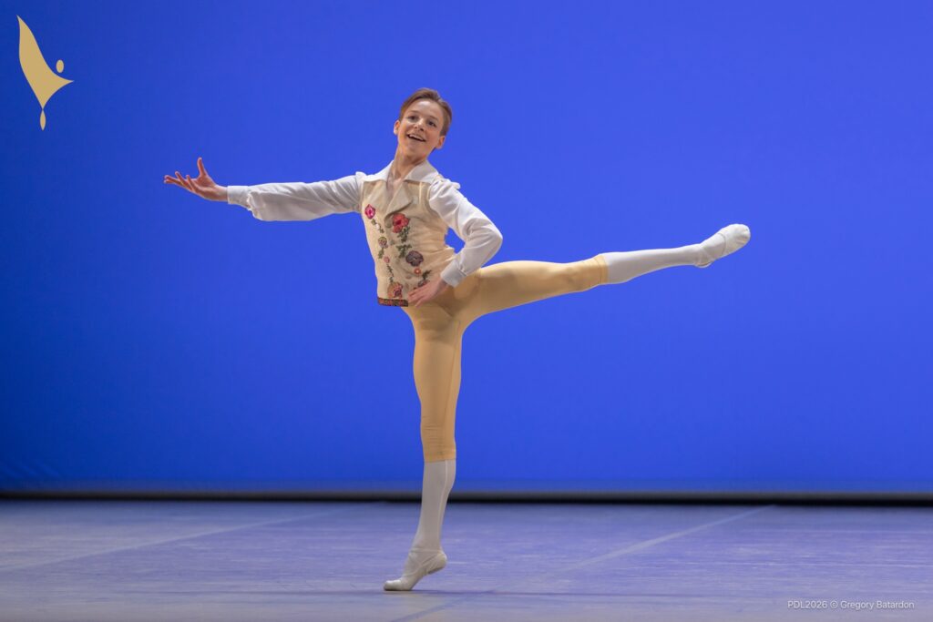 A young dancer does an arabesque with his left hand on his hip and his right arm reaching out to the side. He looks out to the audience with a big smile. He wears a white top and a flowered vest, yellow knickers and white tights.