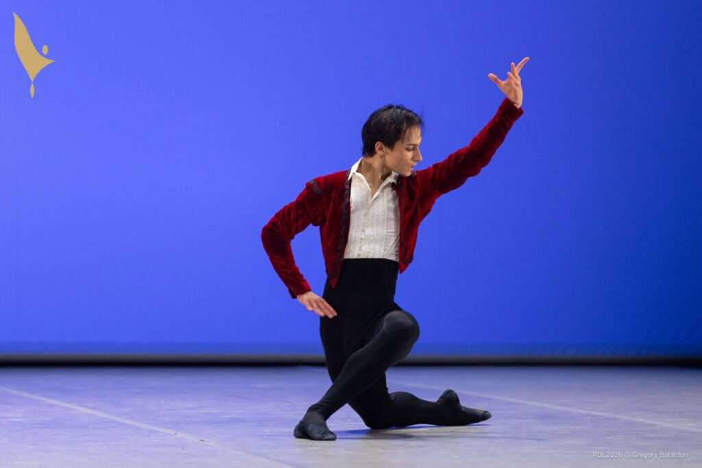 A teenage ballet dancer kneels on his right knee in croisé, with his right hand on his hip and his left arm raised. He turns his head sharply left, and is costumed in a red velvet bolero jacket, white shirt, and high-waisted black tights and shoes.