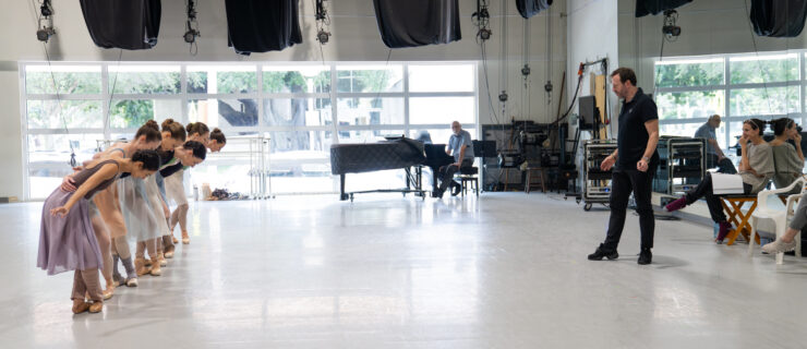 In a large ballet studio, a choreographer dressed in all black faces a line of dancers in front of him, his back to the mirror. The dancers wrap their arms around each others' backs and plié in fifth position, bowing forward.