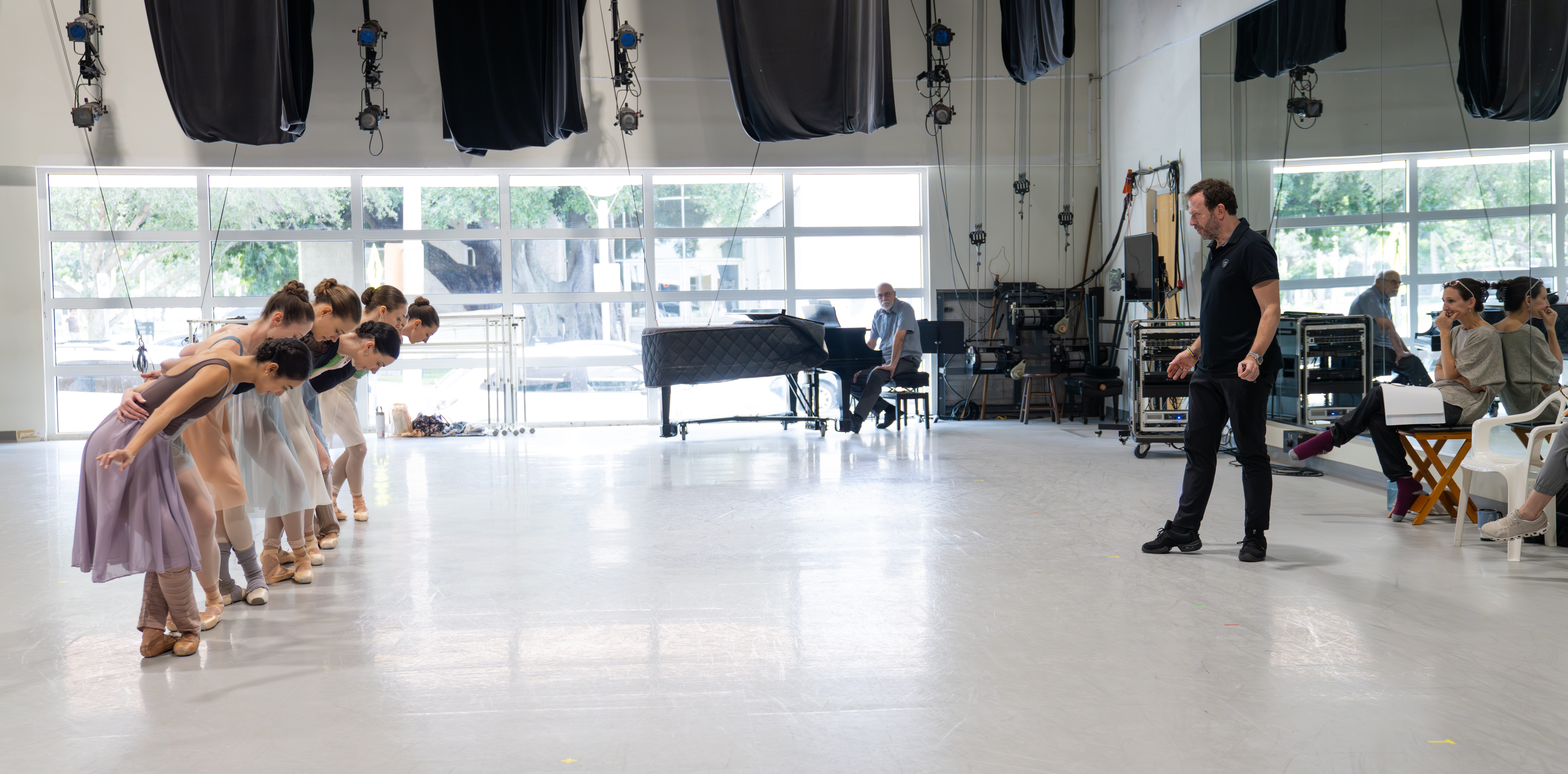 In a large ballet studio, a choreographer dressed in all black faces a line of dancers in front of him, his back to the mirror. The dancers wrap their arms around each others' backs and plié in fifth position, bowing forward.