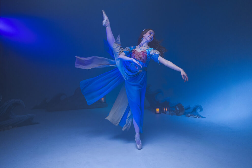 In front of a blue backdrop with an underwater effect, a ballet dancer battements one leg to the side while on pointe, reaching her arms gently toward the opposite direction. She wears a long blue dress with puffy sleeves and red and gold detailing. Her long red hair floats behind her.