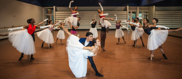 In a large ballet studio, a group of dancers pose in a tableau. Six pairs of partners stand in supported arabesques in two mirrored diagonals, three dancers stand in tendu behind them, and two partners pose in shoulder sits in between. In front, a last pair does a fish pose. The women wear long white skirts.