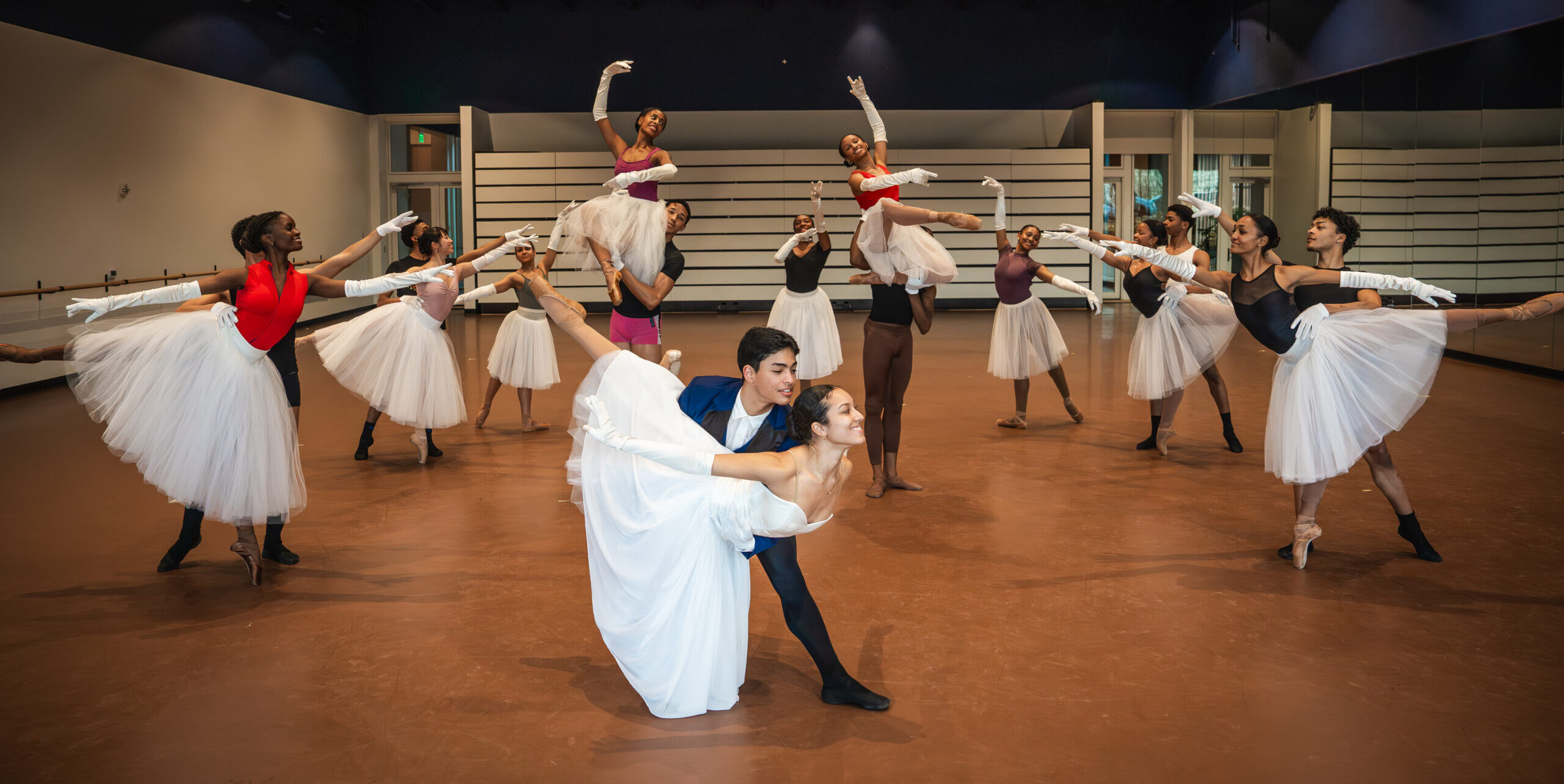 In a large ballet studio, a group of dancers pose in a tableau. Six pairs of partners stand in supported arabesques in two mirrored diagonals, three dancers stand in tendu behind them, and two partners pose in shoulder sits in between. In front, a last pair does a fish pose. The women wear long white skirts.