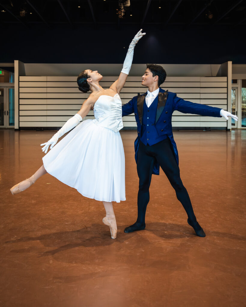 In a ballet studio, two partners rehearse in costume. The woman wears a white dress and gloves and stands on pointe in a low arabesque with one arm reaching high. Her partner, in a dark blue tuxedo, supports her around the waist while standing with one leg propped to the side, his other arm extended.