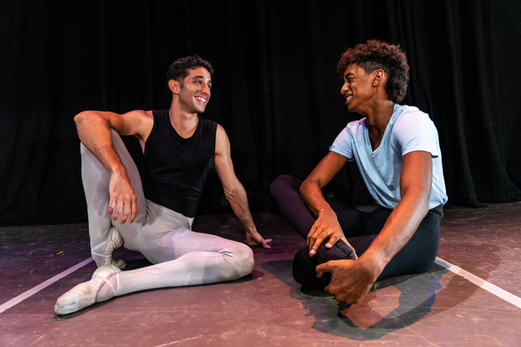 Two male dancers sit on the floor of a stage wearing practice clothes. They smile and have a friendly conversation.