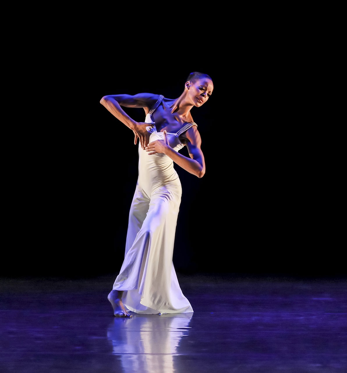 Myrna Kamara, wearing a white gown, stands with her knees bent and leans slightly to her left during a performance. She brings both hands in towards her chest and wears a peaceful expression on her face.