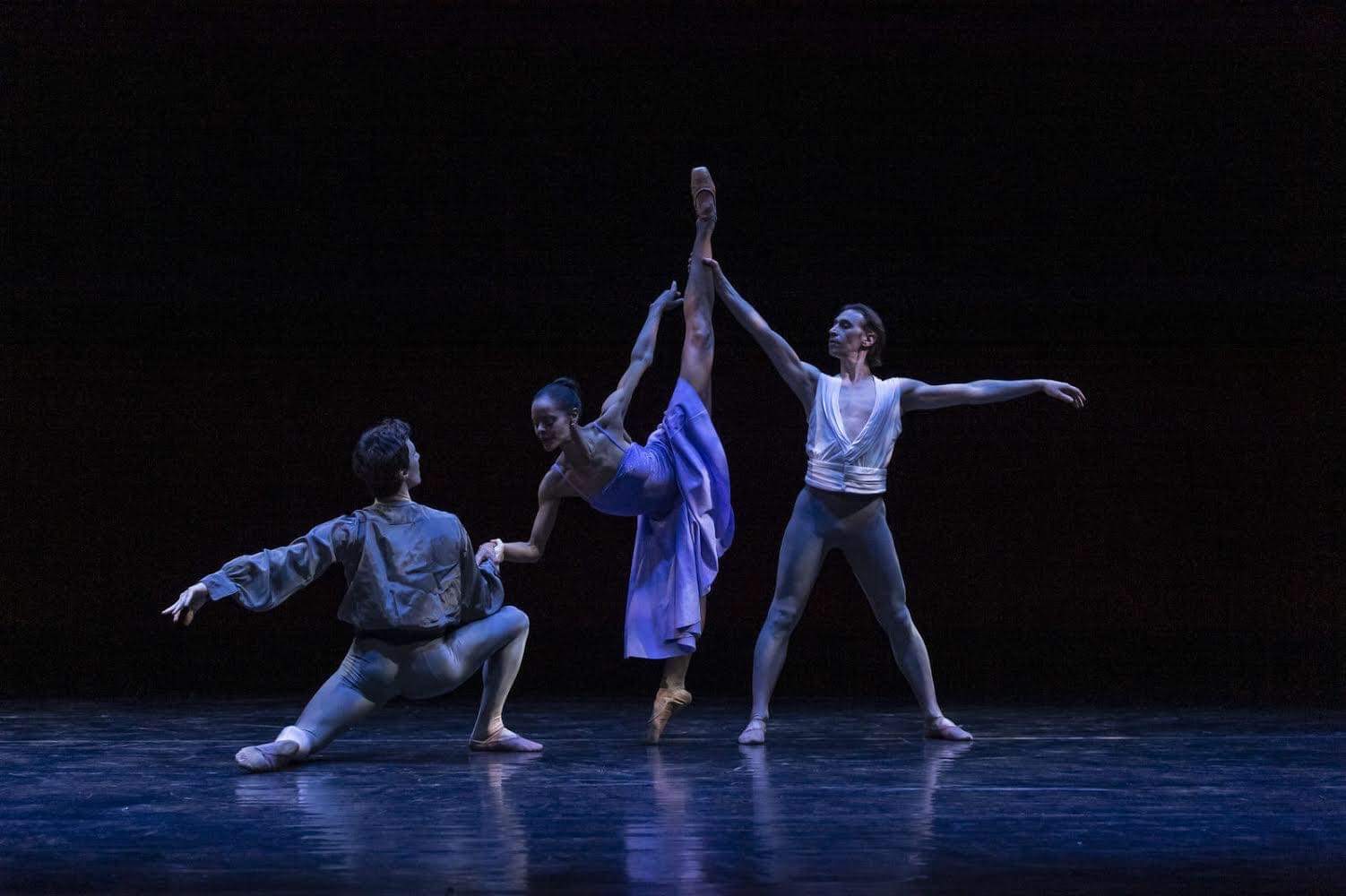 Two men partner a ballerina as she does a penché arabesque. The man on the left wears gray tights and a billowy shirt and kneels in front of her, holding her right hands with his right hand. The other man, in a sleeveless white shirt and gray tights, stands behind her in a wide stance and holds her lifted left leg. The ballerina wears a purple dress, and the stage is dimly lit.
