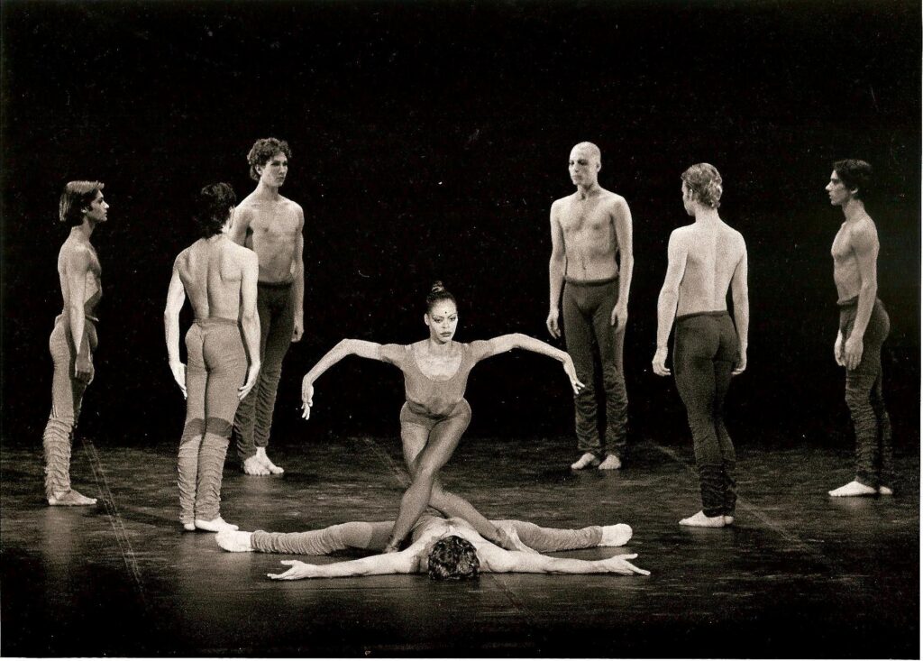Myrna Kamara stands over a man lying onstage, her legs bent and in b-plus while she holds her arms out to thte side, curving them at the elbow. Six shirtless men in pants stand form a circle behind her, facing each other and standing in sixth position.