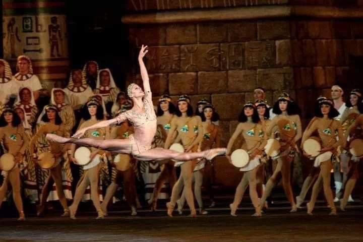 Myrna Kamara does a saut de chat onstage in front of a large corps de ballet. She wears a sparkly, gold cropped top with matching briefs and headpiece. The dancers behind her wear dark shoulder-length wigs and flesh-colored unitards, and stand in their right legs holding a drum against their right hip.